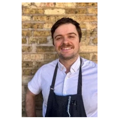 Smiling man in white shirt and dark apron standing against a brick wall.