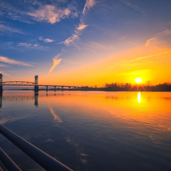 Sunset over a calm river with a bridge and boardwalk.