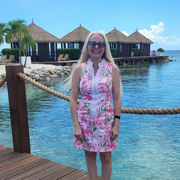 A woman in a floral dress stands on a pier with thatched huts and clear water behind her.