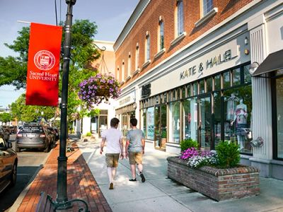 Two people walk on a sidewalk near a boutique and a university banner.
