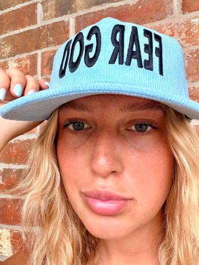 Woman wearing a light blue cap with 'FEAR GOD' text, posing against a brick wall.