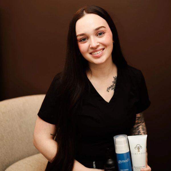 Smiling woman with long dark hair holding skincare products, sitting in a beige chair.