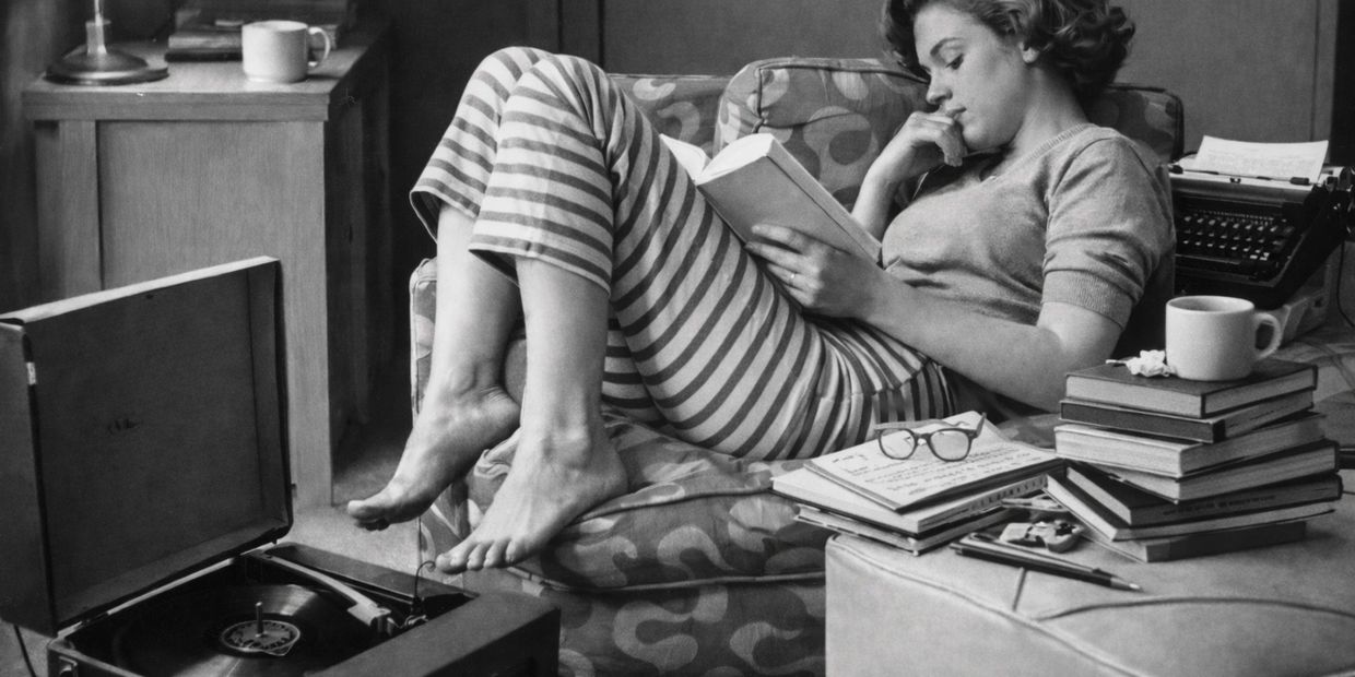 Mulher deitada em uma poltrona lê um livro em um ambiente doméstico, cercada por livros e um toca-discos, em fotografia em preto e branco.