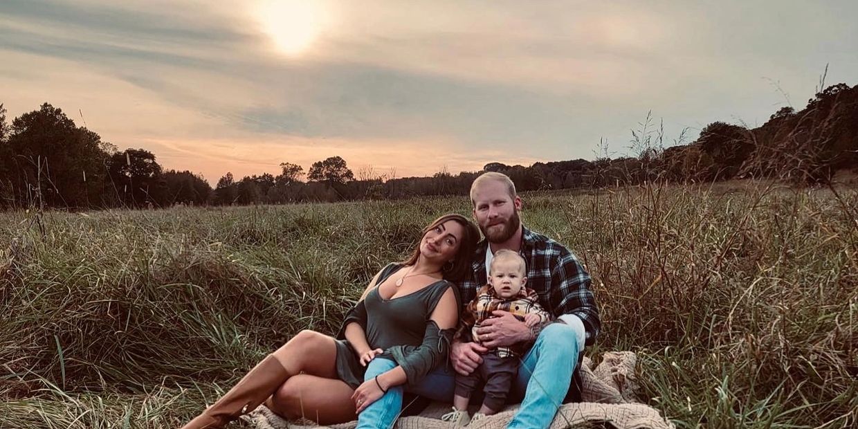 A family of three sits on a blanket in a grassy field at sunset.