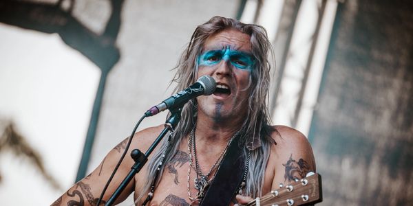 A tattooed male musician with grey hair sings into a mic while playing an acoustic guitar on stage.