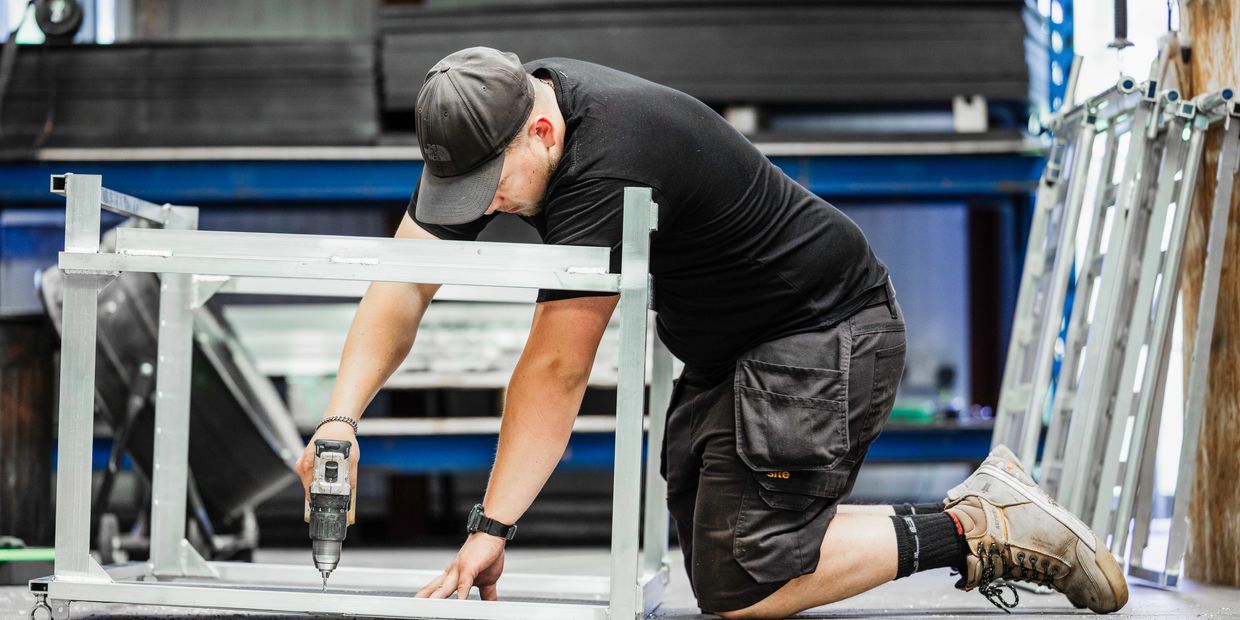 A tradesman wearing workwear uses a cordless drill to assemble a metal frame inside a workshop.