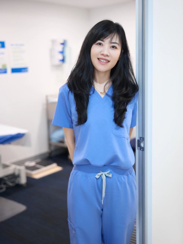 A smiling nurse in blue scrubs stands in a clinical room doorway.