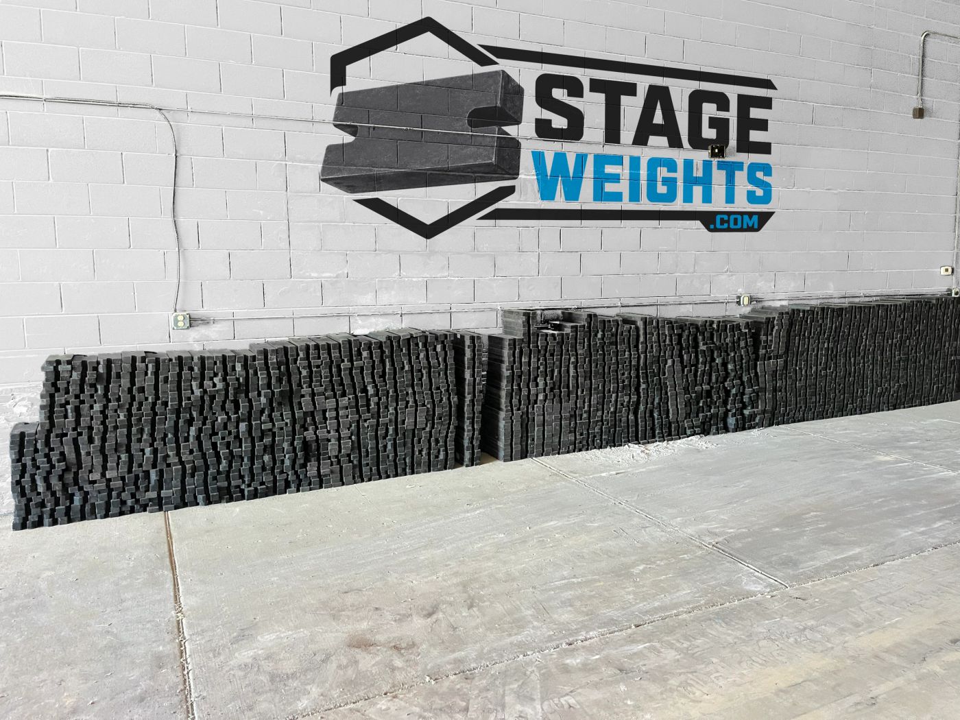 Long rows of black steel stage weights stacked against a white warehouse wall painted with the Stage Weights logo