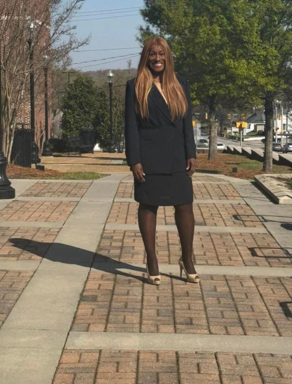 A woman in a black suit and beige heels stands on a sunny brick pathway.