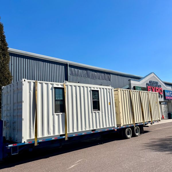 Two large shipping containers with windows mounted on a flatbed trailer under a clear blue sky.