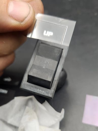 Hand holding a black switch with a protective film labeled 'UP'.