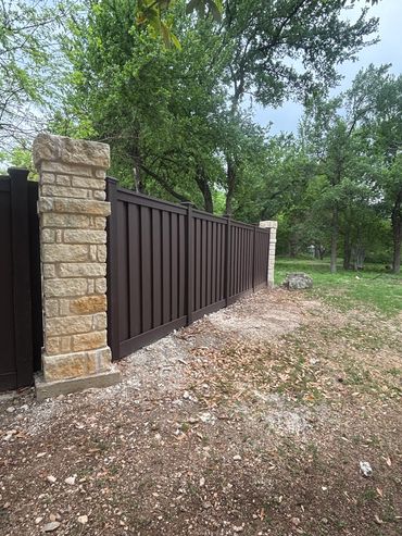 Brown fence with stone pillars, set against a green, leafy backdrop.