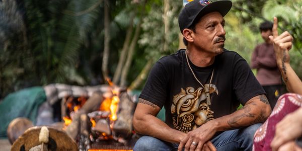 Man in black shirt sitting near a fire in a forest setting.
