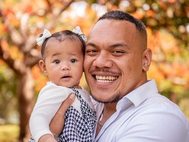 A smiling man holding a baby girl outdoors with autumn leaves in the background.