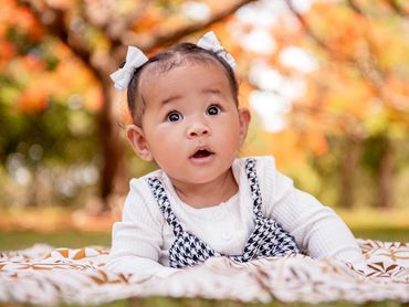 Surprised baby with bows in hair lying on blanket outdoors in autumn.