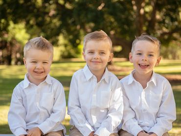 Three young boys in white shirts sitting outdoors, smiling.