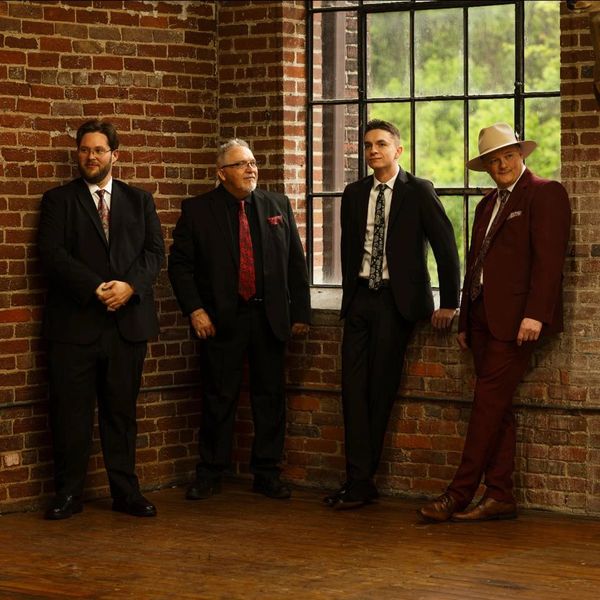 Four men in suits standing against a brick wall near a large window.