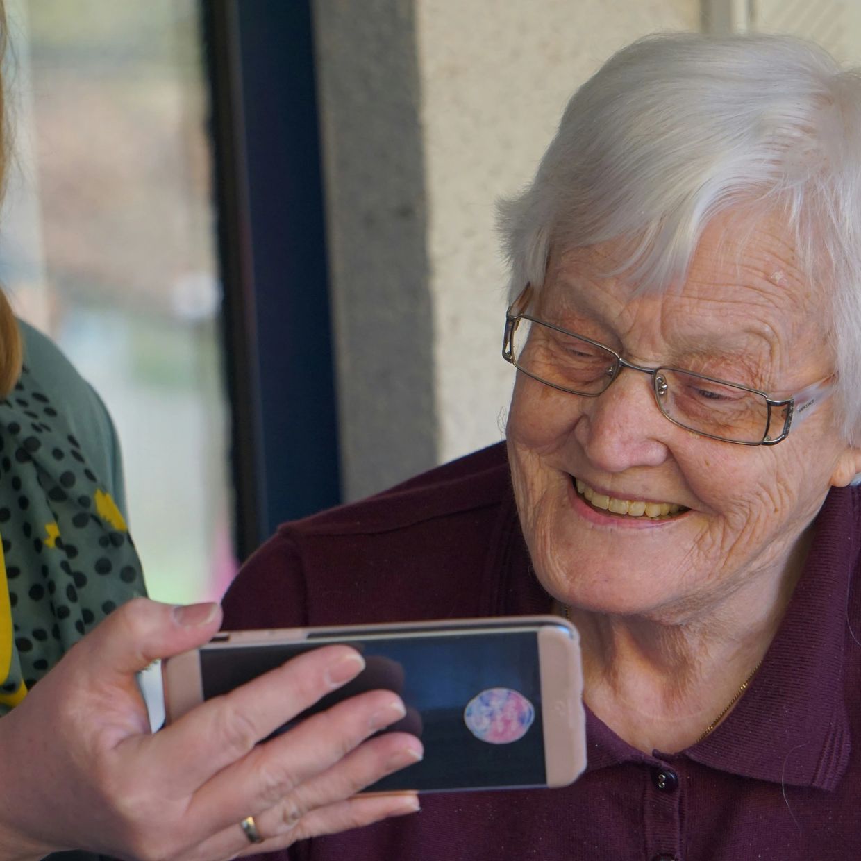 Elderly woman smiling while looking at a phone held by a masked caregiver.