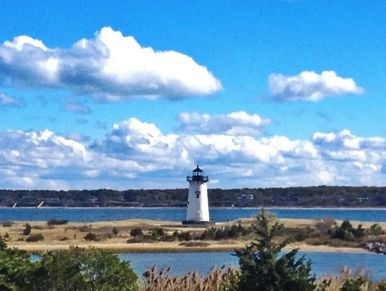 A lighthouse stands near the coast under a blue sky with scattered clouds.