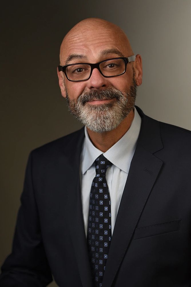 Smiling bald man with glasses in a suit and patterned tie.