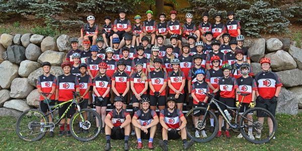 Large youth cycling team posing outdoors with helmets and bikes.