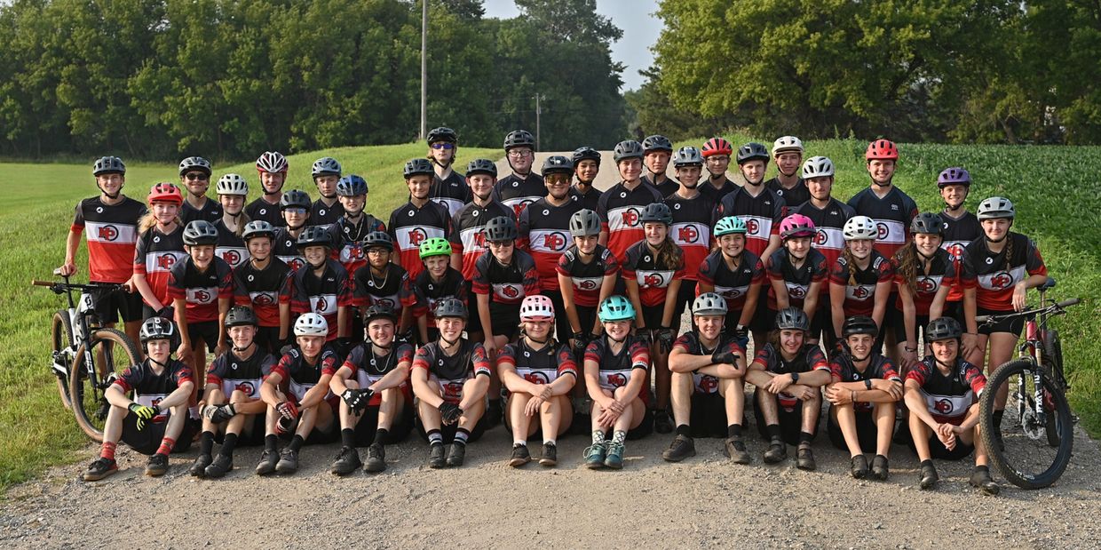 Large youth cycling team posing outdoors in matching jerseys and helmets.