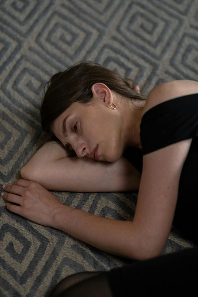 A woman lies on a patterned carpet, looking pensive and tired.