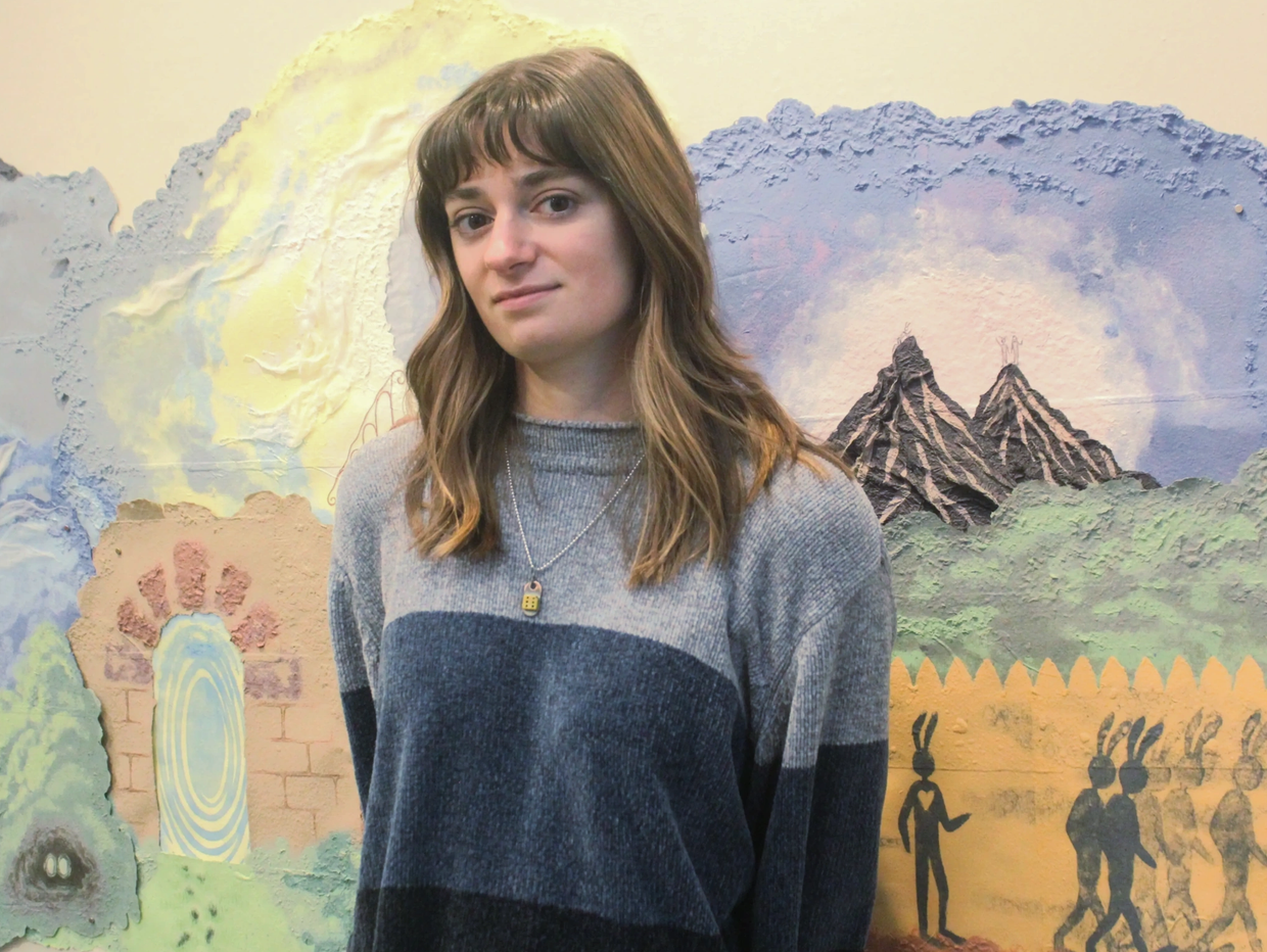 A woman stands in front of a colorful mural with mountains and rabbits.