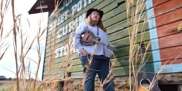 Zach stands with his mandolin against a building. There is tall oat grass in the foreground