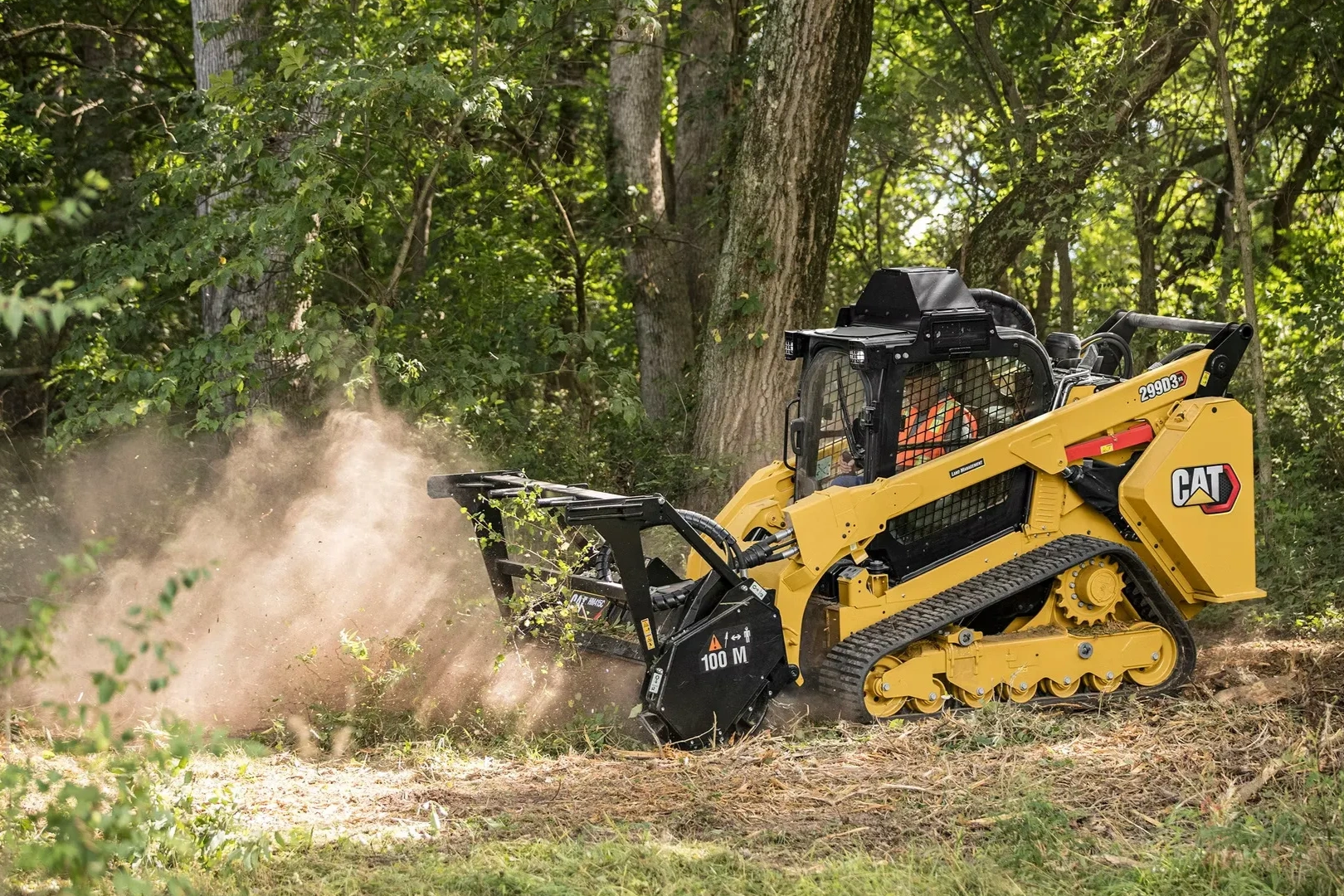 A yellow CAT compact track loader clearing brush in a forested area.