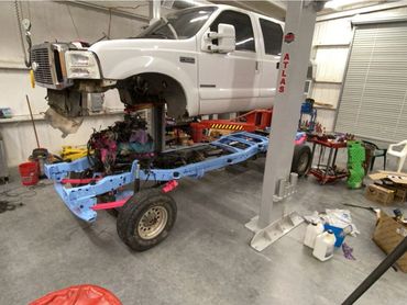 A white truck lifted on a hydraulic lift with its chassis painted blue and pink in a garage.