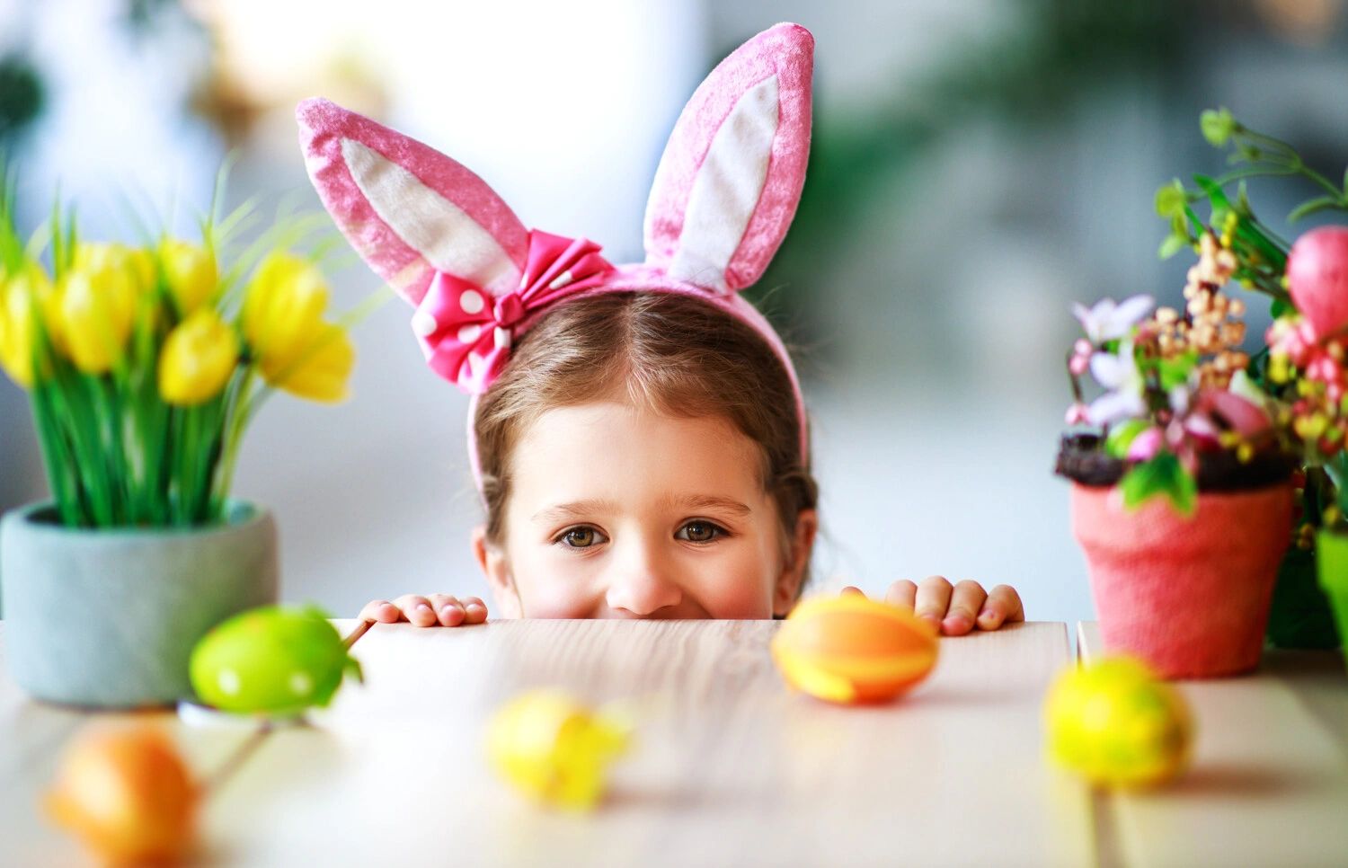 A child wearing pink bunny ears peeks over a table with Easter decorations.