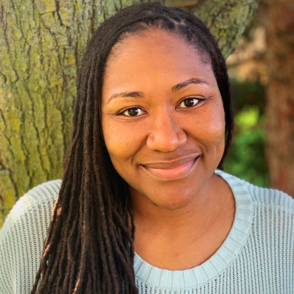 Person with long locs, wearing a light sweater, smiling while standing outdoors near a tree.



