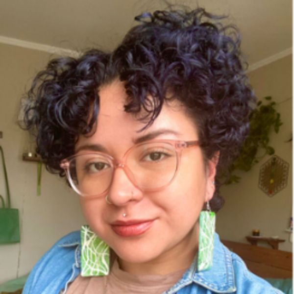 Person with curly hair, glasses, and green earrings, wearing a denim shirt and smiling at the camera.
