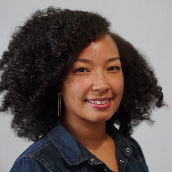 Person with curly hair smiling, wearing a denim shirt and earrings standing against white background