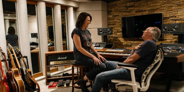 Two people laughing in a music studio filled with guitars and equipment.