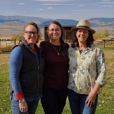 Three women smiling outdoors with mountains in the background.
