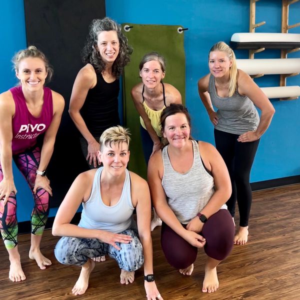 Six smiling women in workout clothes posing barefoot in a fitness studio.