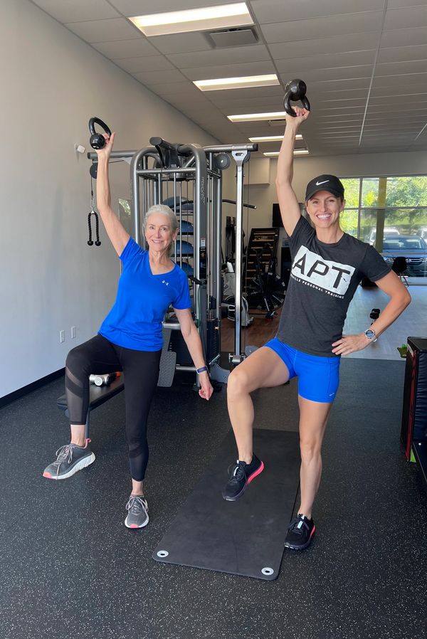 Two women exercising with kettlebells in a gym, smiling and lifting one leg.