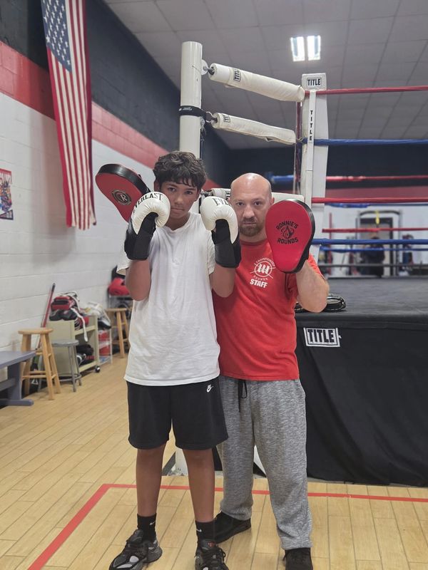 A young boxer and coach pose with boxing gloves in a gym.