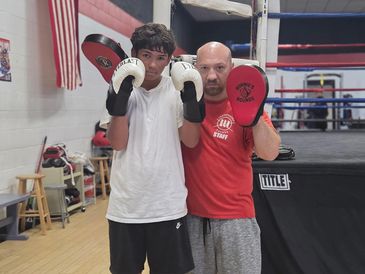 A young boxer and coach pose with boxing gloves in a gym.