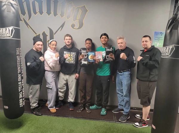 A group of seven people posing with boxing gloves and books in a gym.