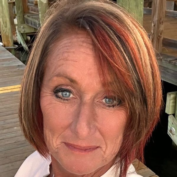 Close-up of a woman with blue eyes and highlighted hair on a wooden dock.