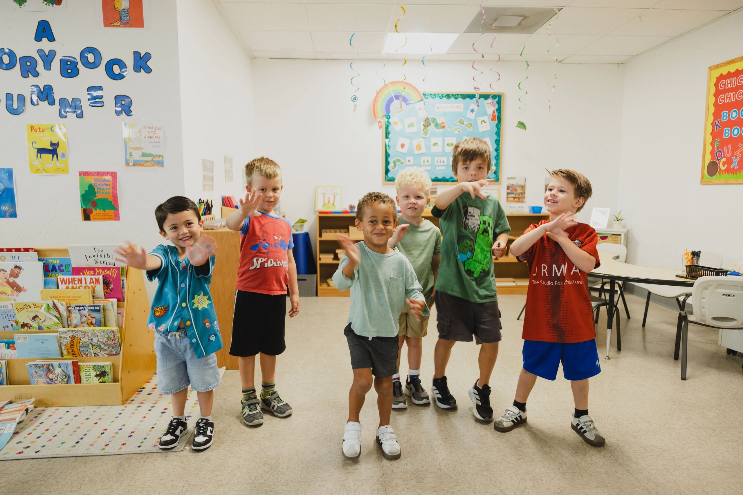 Six young boys waving and smiling in a colorful classroom.