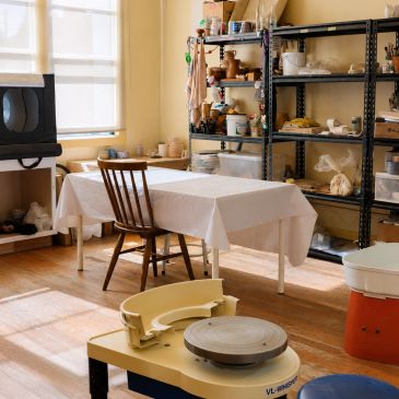 A pottery studio with a wheel, shelves, and a table covered in a white cloth.