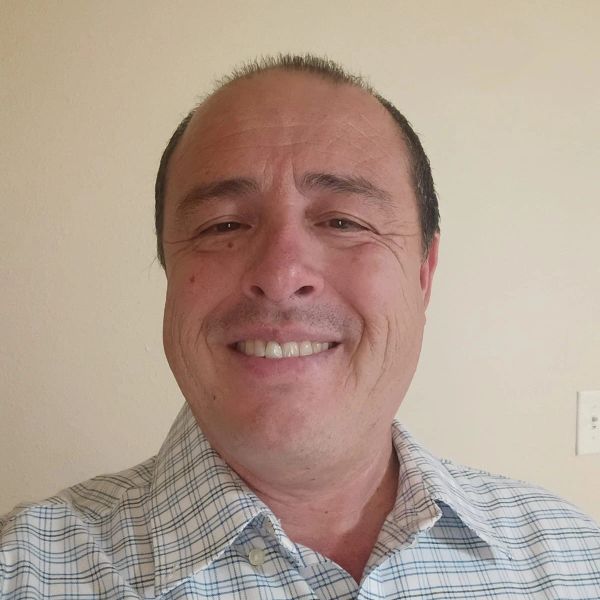 Smiling man wearing a checked shirt standing against a plain wall.