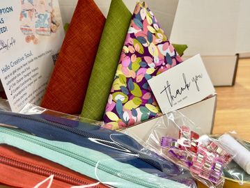 A sewing kit with colorful fabrics, zippers, clips, thread, and a thank you card.