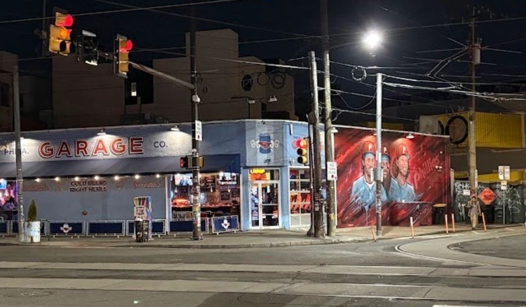 Night view of a corner bar with a mural of three people on its side wall.