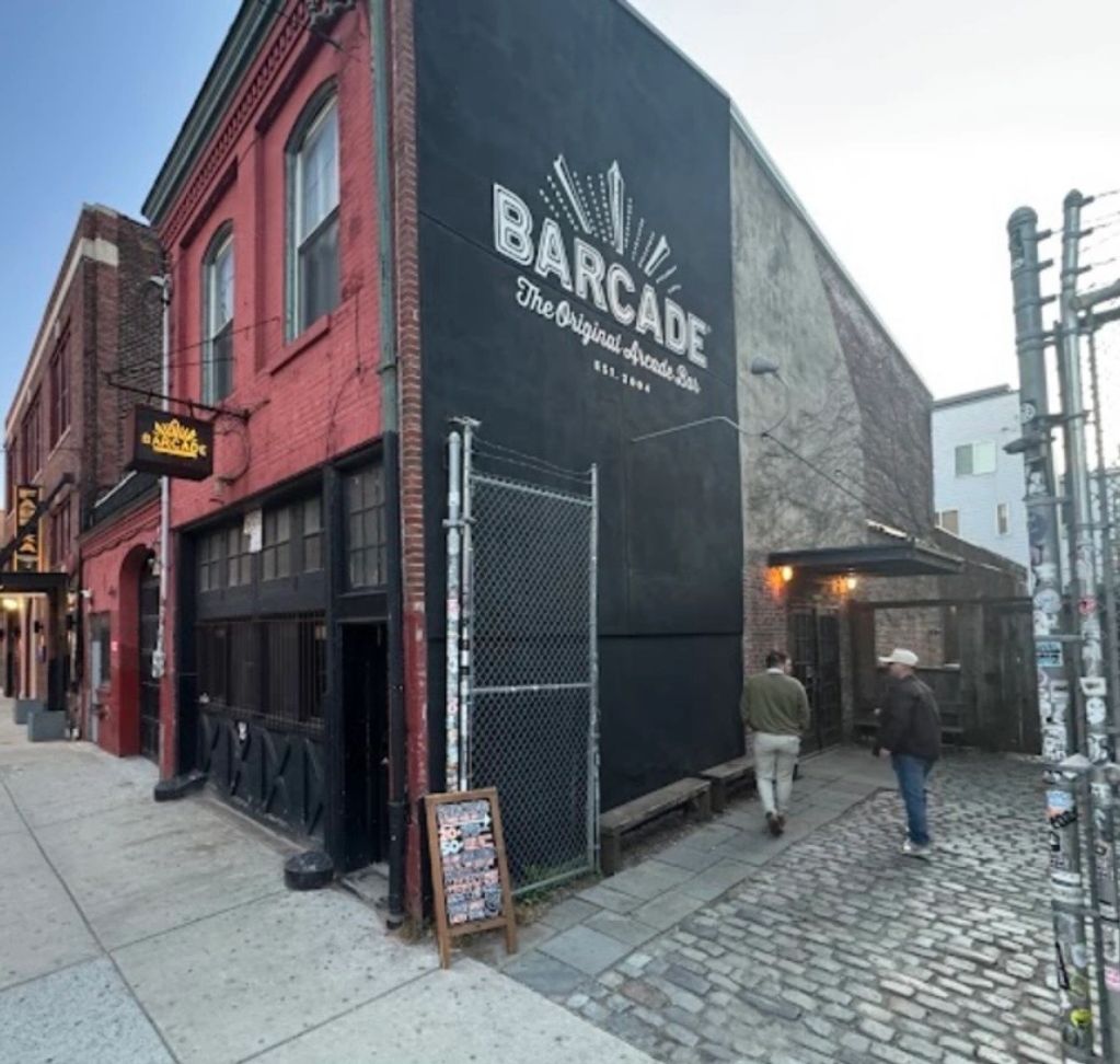 Two men walk past Barcade, an arcade bar in a brick building on a cobblestone street.