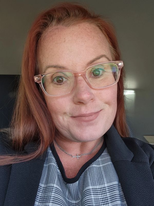 Smiling woman with red hair and glasses wearing a plaid top and black jacket.